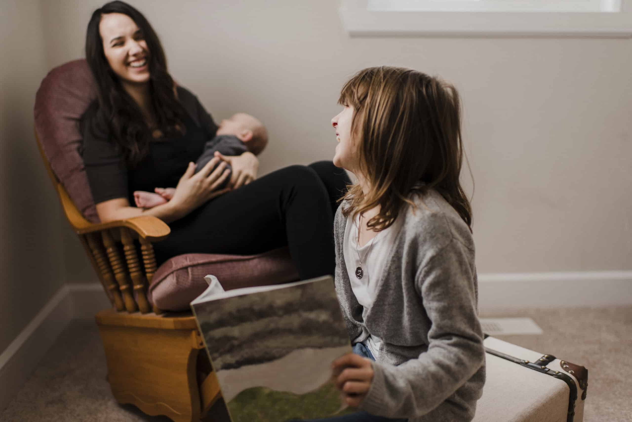 girl laughing with mother while she cradles baby brother in living room armchair